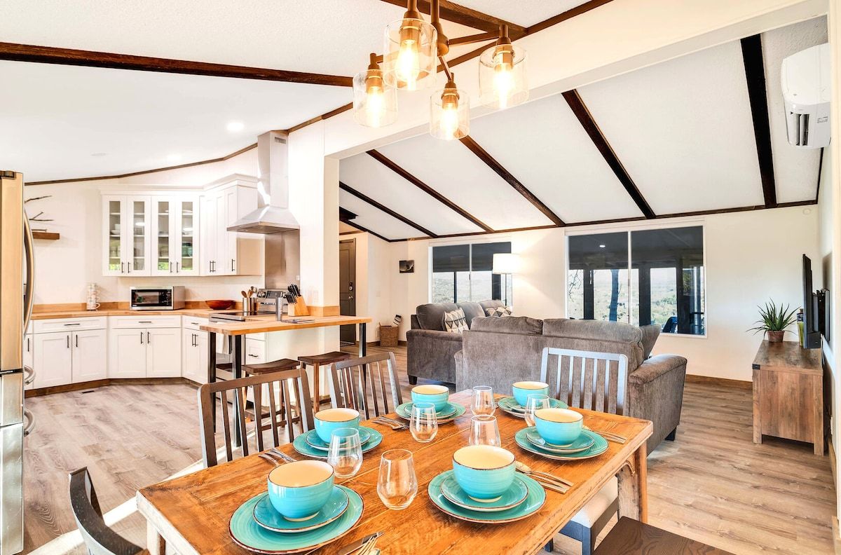 Une cuisine spacieuse avec des meubles blancs, un coin repas avec une table en bois dressée pour quatre personnes, et un salon avec un canapé et une grande fenêtre.