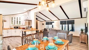 Une cuisine spacieuse avec des meubles blancs, un coin repas avec une table en bois dressée pour quatre personnes, et un salon avec un canapé et une grande fenêtre.