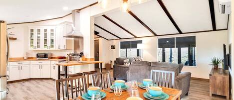 A spacious kitchen with white cabinets, a dining area with a wooden table set for four, and a living area with a sofa and a large window.