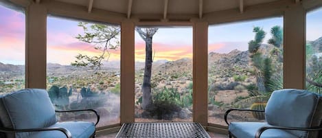 Una habitación con dos sillas y una mesa, amplias ventanas con vista al desierto y techo de madera.