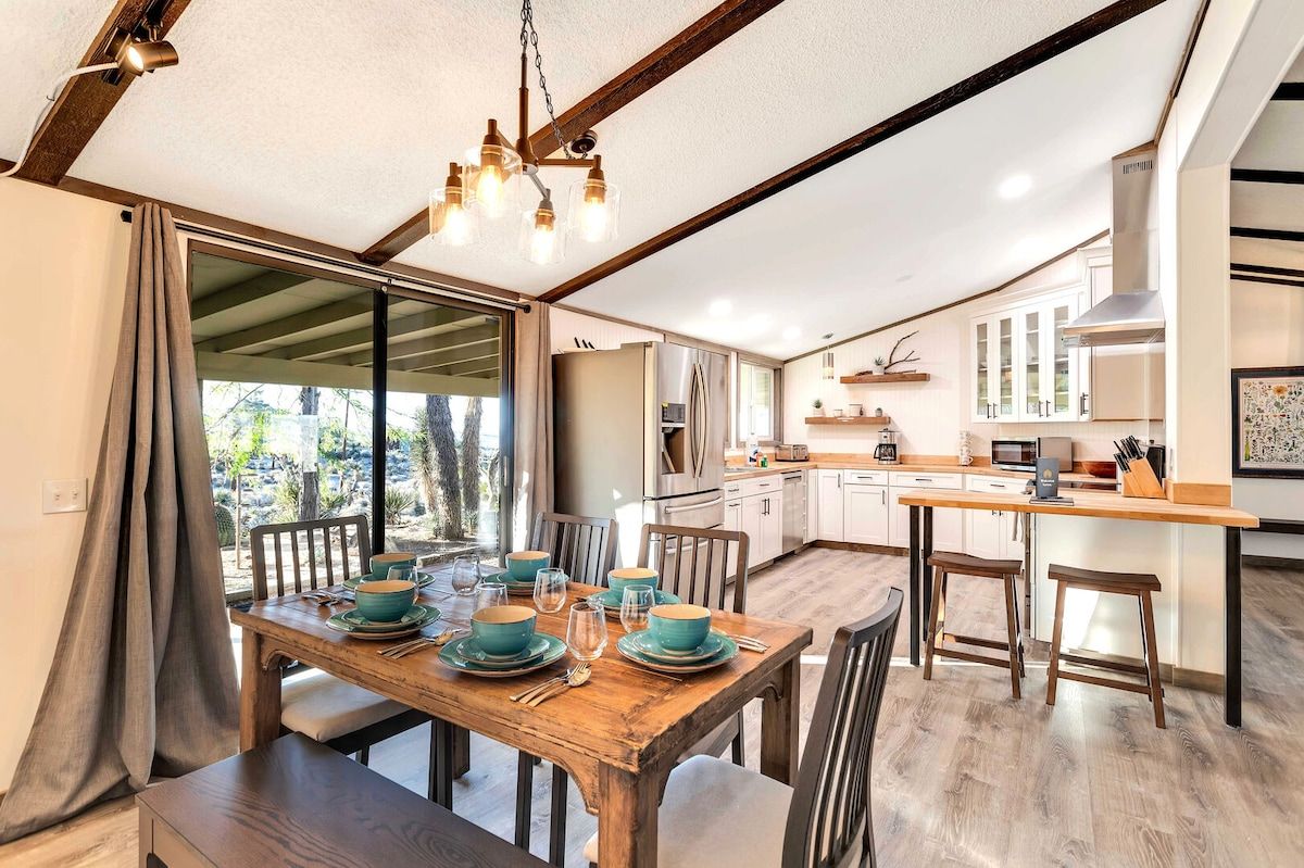 Un comedor con una mesa de madera dispuesta para cuatro personas, una cocina con armarios blancos y un ventanal con vistas al exterior.