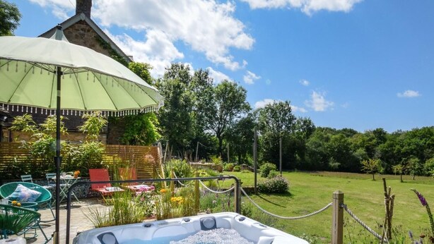 Ein Whirlpool in einem Garten mit einer Holzterrasse, Gartenmöbeln und einem Steinhaus im Hintergrund.