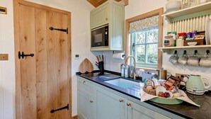 A kitchen with wooden cabinets, a microwave, a sink, and a window with a view of greenery. - 1 Bed in Tewkesbury (oc-94686) (Bushley)