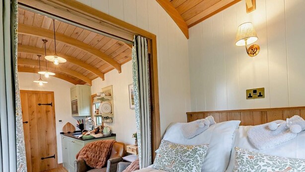 A cozy bedroom with a bed, pillows, and a nightstand. There is a wooden ceiling and a wall-mounted light fixture. - 1 Bed in Tewkesbury (oc-94686) (Bushley)