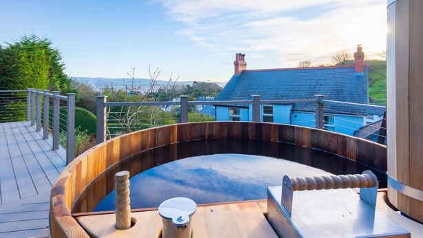 A wooden hot tub on a deck with a view of a house and hills. - 7 Bed in Aberdyfi (oc-dy030) (Aberdovey)
