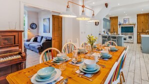 A dining area with a long wooden table set for a meal, a piano, and a kitchen in the background. - 7 Bed in Aberdyfi (oc-dy030) (Aberdovey)
