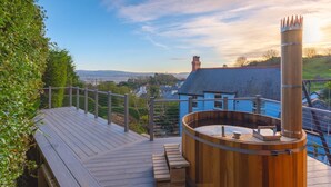 A wooden deck with a hot tub, a metal railing, and a view of a house and landscape. - 7 Bed in Aberdyfi (oc-dy030) (Aberdovey)
