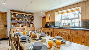 A kitchen with wooden cabinets, a dining table set with plates, bowls, and glasses, and a window with a view. - 4 Bed in Bude (oc-41608) (Poundstock)