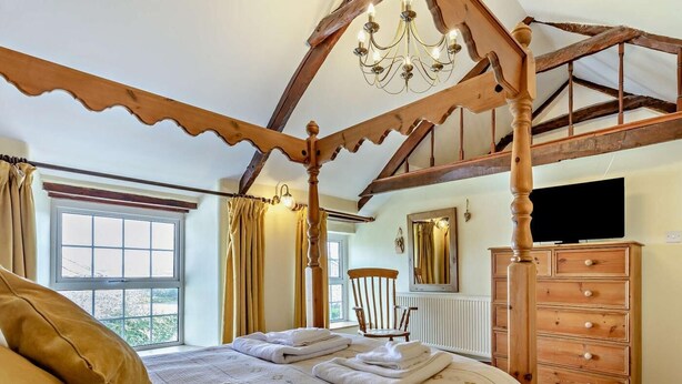 A bedroom with a four-poster bed, a wooden dresser, a chair, a television, and a window with curtains. - 4 Bed in Bude (oc-41608) (Poundstock)