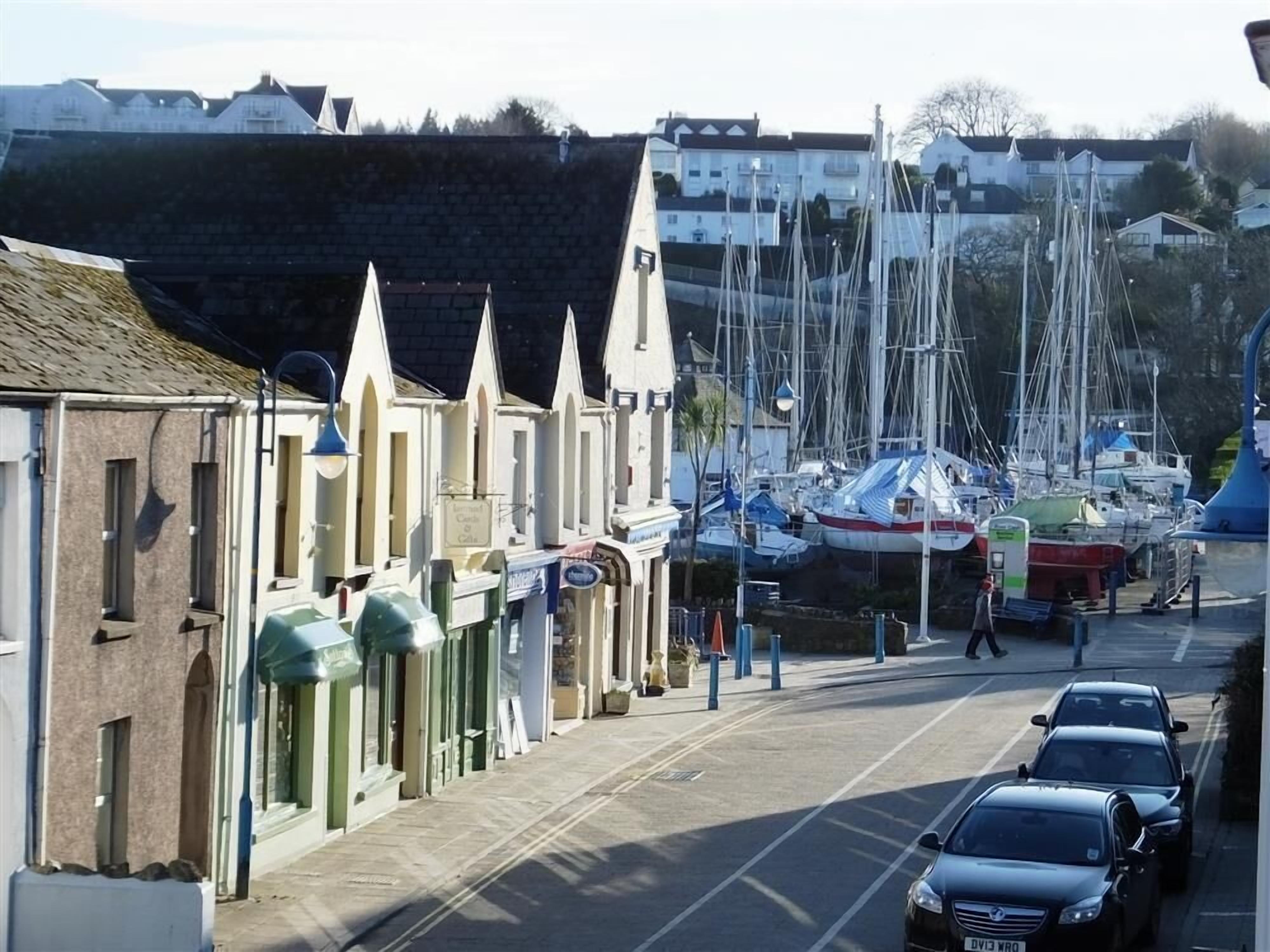 Une rue bordée de voitures garées, une rangée de bâtiments aux toits en pente et un port de plaisance avec des bateaux en arrière-plan.