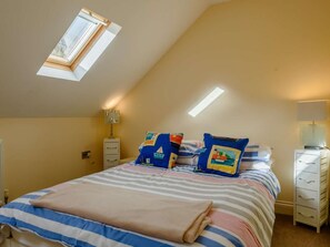 Ein Schlafzimmer mit schrägen Wänden, einem Oberlicht, einem Bett mit gestreifter Bettwäsche und einem Nachttisch mit Lampe.