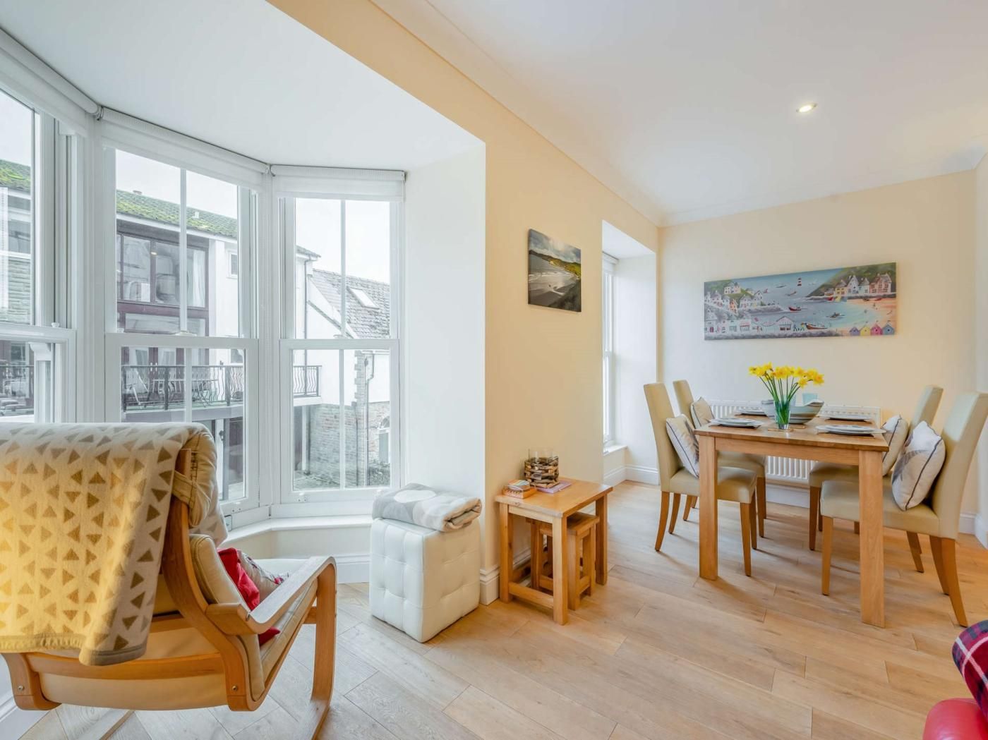 A bright living room with a large window, a dining area with a table and chairs, and a cozy seating area with a bench and cushions.