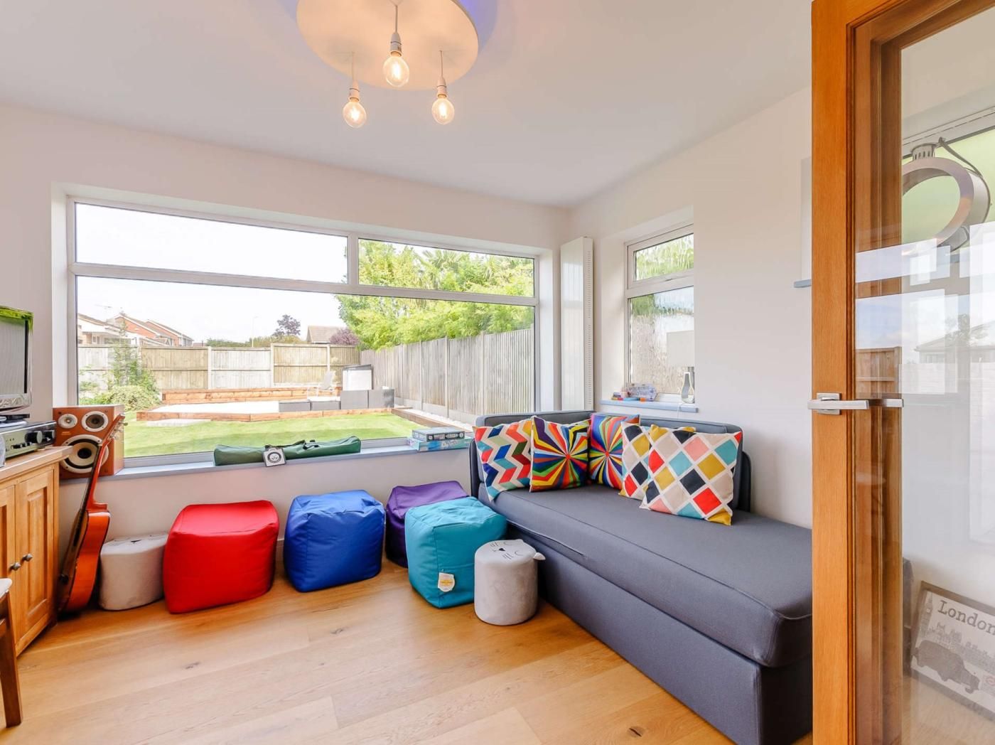 Ein Zimmer mit grauem Sofa, bunten Kissen, einem Holzschrank und einem Fenster mit Blick auf einen Garten.