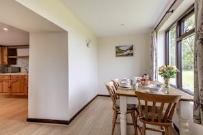 A dining area with a wooden table set for four, a kitchen in the background, and a window with curtains. - 2 Bed in Woolacombe (oc-43279) (Woolacombe)