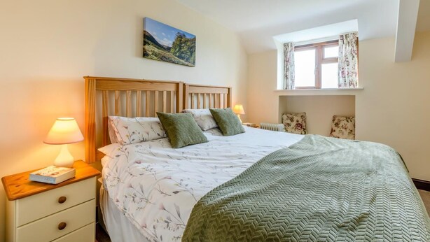 A bedroom with a wooden headboard, a bed with floral bedding, a green throw blanket, a nightstand with a lamp, and a window with curtains. - 2 Bed in Woolacombe (oc-43279) (Woolacombe)
