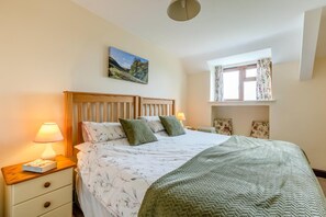 A bedroom with a wooden headboard, a bed with floral bedding, a green throw blanket, a nightstand with a lamp, and a window with curtains. - 2 Bed in Woolacombe (oc-43279) (Woolacombe)