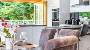 Moderni keittiö, jossa on suuri ikkuna, ruokailutila kahdelle katetulla pöydällä sekä näkymä vehreään maisemaan.