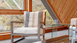 Ein gemütliches Zimmer mit einem raumhohen Holzrahmenfenster, einem gestreiften Sessel, einem Glastisch und Blick auf Bäume.