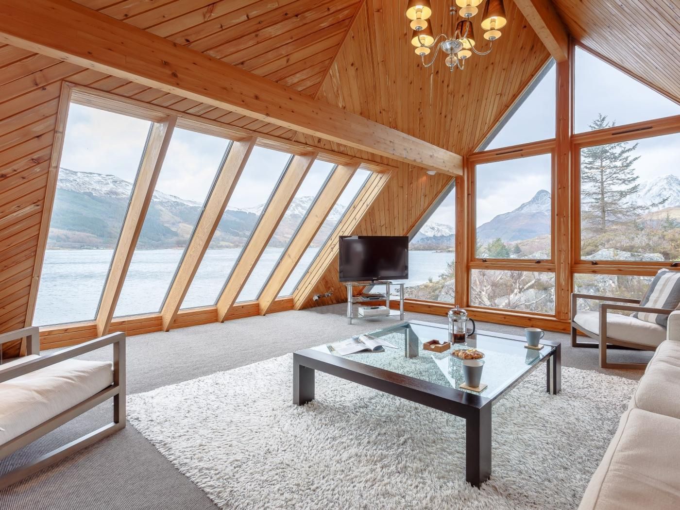 A modern interior with a large window offering a view of a lake and mountains, a flat-screen TV, and a cozy seating area.