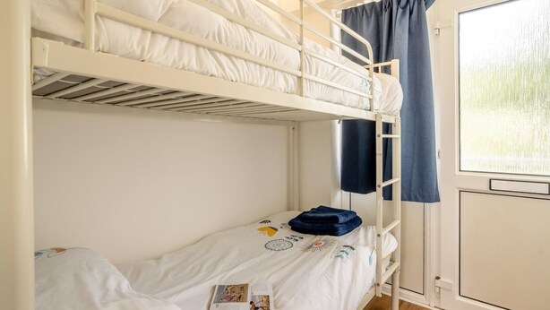 Une chambre avec un lit superposé, une fenêtre, une porte et une petite table de chevet sur laquelle se trouve un livre.