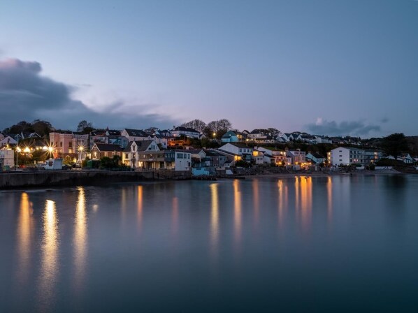 Uma vila costeira ao entardecer, com edifícios ao longo da orla, água calma refletindo as luzes e céu claro.