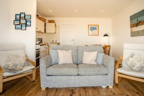 A living room with a grey sofa, two armchairs, a wooden floor, a kitchen in the background, and a wall clock. - 1 Bed in Saundersfoot (oc-fb243) (Saundersfoot)