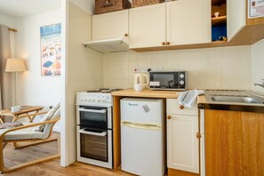 A compact kitchen with a stove, oven, microwave, and refrigerator. There is a wooden table with a chair and a lamp. - 1 Bed in Saundersfoot (oc-fb243) (Saundersfoot)