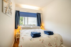 Quarto com cama, almofadas, mesa de cabeceira, abajur e janela com cortinas.