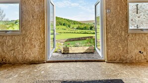 Een kamer met een groot raam dat uitzicht biedt op een groen landschap, een klein houten bankje en een lijst met een afbeelding aan de muur.