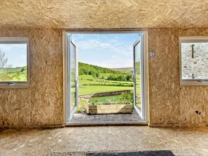 Ein Zimmer mit großem Fenster, das Blick auf eine grüne Landschaft freigibt, eine kleine Holzbank und ein gerahmtes Bild an der Wand.