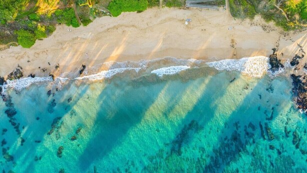 Vista aérea de una playa con aguas turquesas cristalinas, orilla de arena y algo de vegetación.