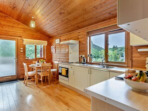 A wooden cabin interior with a dining area, kitchen, and large windows offering a view of the outdoors.