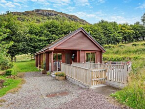 A small wooden cabin with a porch, surrounded by a gravel driveway and a wooden fence.