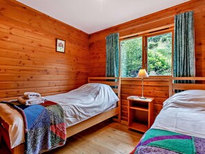 A cabin room with two wooden beds, a window with curtains, a bedside table with a lamp, and a framed picture on the wall.