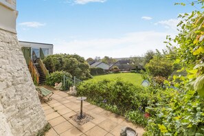 A stone wall, a patio with a table and chairs, a garden with a lawn and trees, and a view of neighboring houses. - 6 Bed in Bideford (oc-beaho) (Bideford)