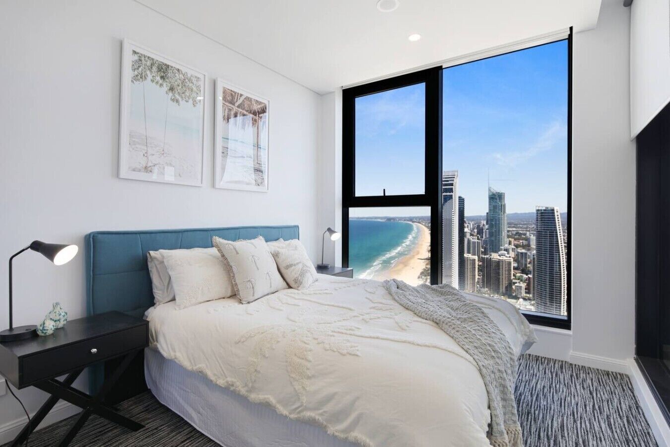 A modern bedroom with a large bed, a nightstand, a lamp, and a view of a beach and cityscape through a large window.