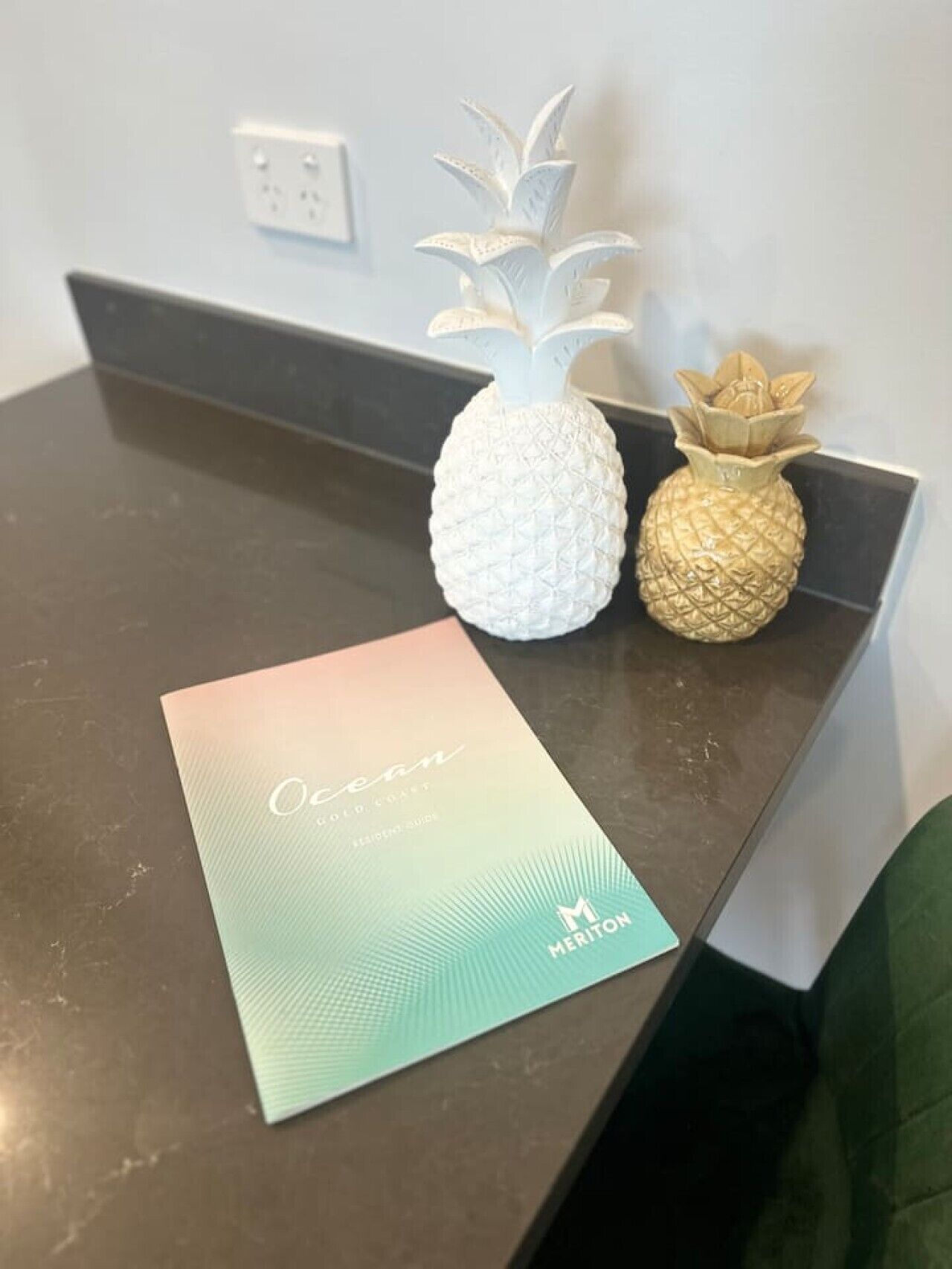 A brochure titled 'Ocean Gold Coast' on a dark countertop with two pineapple-shaped vases, one white and one gold, and a power outlet in the background.