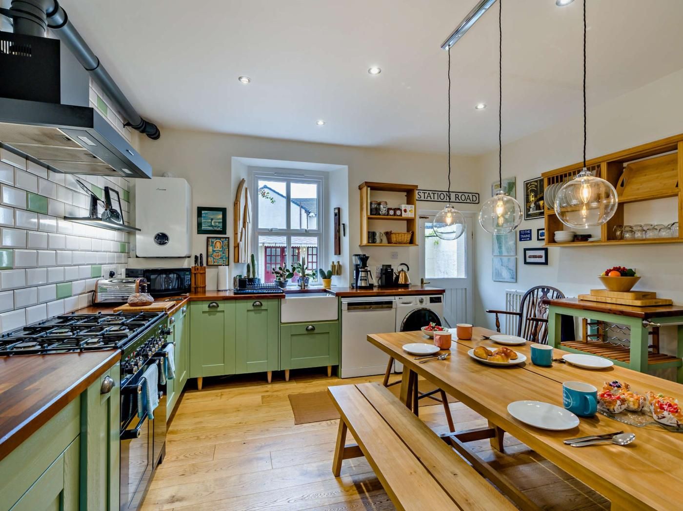 Een keuken met een gasfornuis, groene kastjes, een houten eettafel en hangende pendelverlichting.