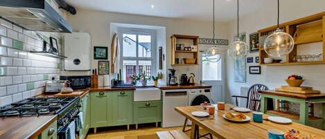 Een keuken met een gasfornuis, groene kastjes, een houten eettafel en hangende pendelverlichting.