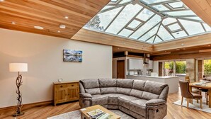 A modern living room with a wooden ceiling, a glass skylight, a grey sectional sofa, a wooden coffee table, and a dining area with a table and chairs. - 1 Bed in Alnwick (oc-94771) (Alnwick)