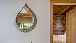 A room with a wooden ceiling, a stone wall, a bed with a floral bedspread, a wooden door, a mirror with a decorative frame, and a rabbit sculpture on a wooden table. - 1 Bed in Alnwick (oc-94771) (Alnwick)