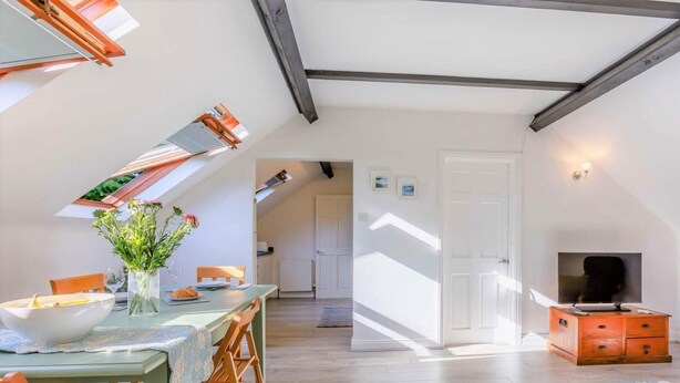Ein Essbereich mit Tisch, Stühlen und einem Blumenstrauß. Im Wohnbereich befindet sich ein Fernseher und ein Schrank. Der Raum hat Holzdielen und eine schräge Decke.