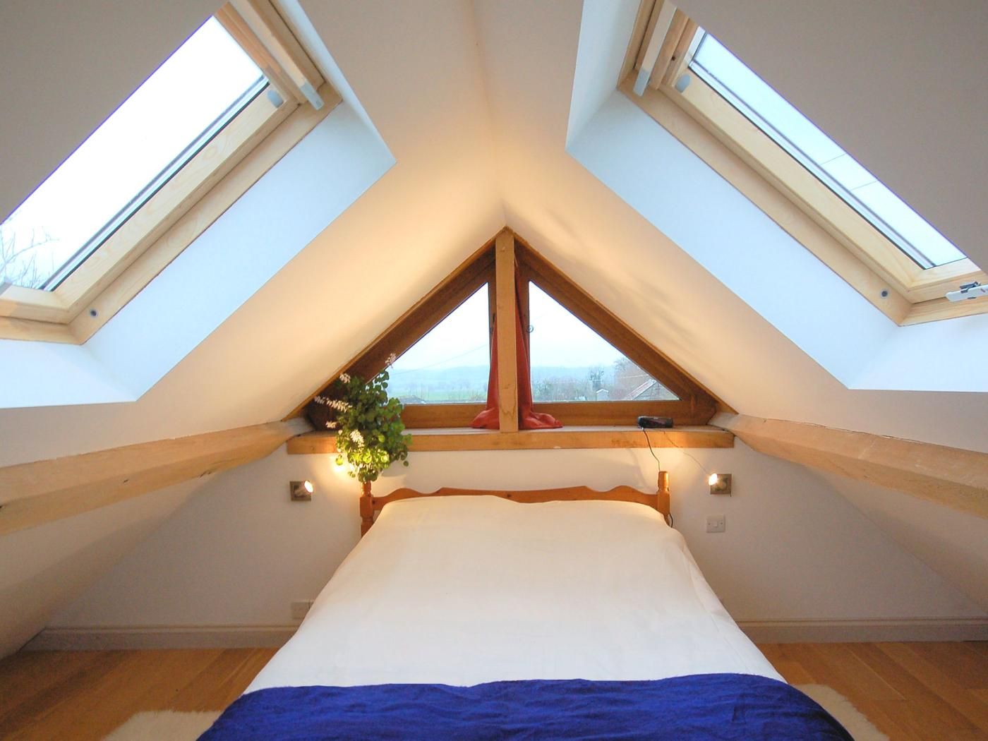 A bedroom with a sloped ceiling and skylights, a bed with a blue blanket, and a window with a plant.