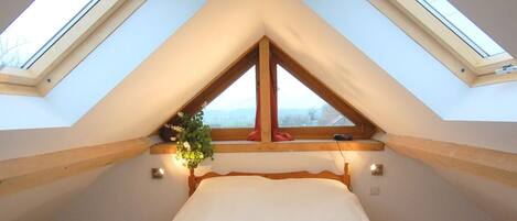A bedroom with a sloped ceiling and skylights, a bed with a blue blanket, and a window with a plant.