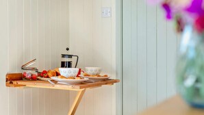 A kitchen area with a wooden table, a coffee maker, a jar of strawberries, and two stools. - 1 Bed in Fowey (oc-95015) (Golant)