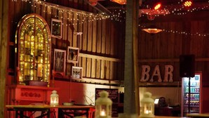 A rustic bar with wooden walls, string lights, and a large arched window. Tables and chairs are arranged for seating. - 1 Bed in Fowey (oc-95015) (Golant)