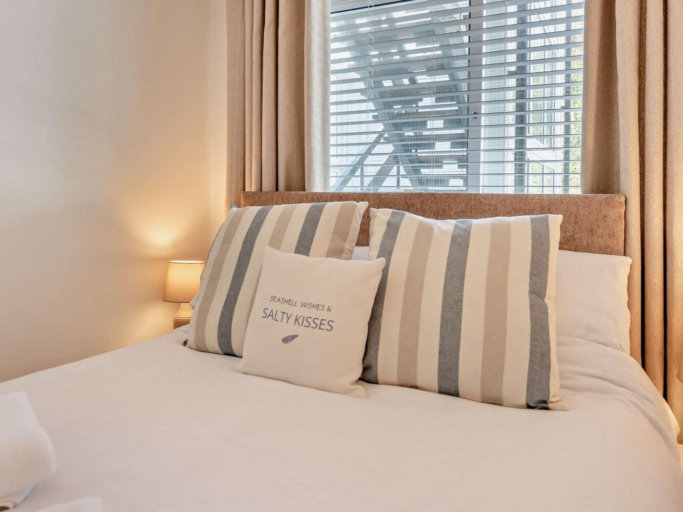 A hotel room with a bed, striped pillows, a window with blinds, and a lamp.