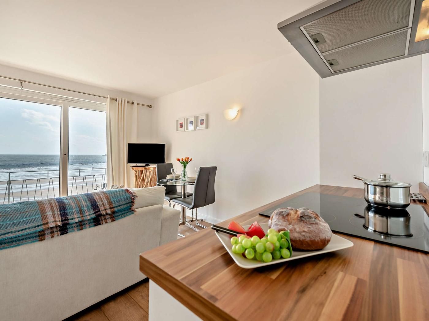 A modern living room with a sofa, a dining table with a plate of fruit, a television, and a view of the sea.
