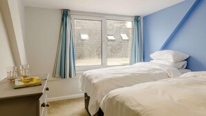 A hotel room with two beds, a desk with glasses and a book, a window with curtains, and a view of a brick building. - 5 Bed in Tenby (oc-83907) (Tenby)
