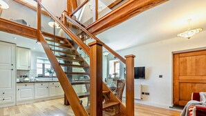 Una escalera de madera con barandillas de vidrio que lleva a una zona de cocina con armarios blancos y una mesa de comedor.
